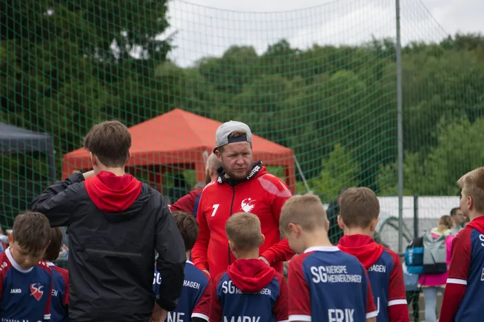 Ein Mann mit Baseballkappe und rotem Trainingsanzug steht inmitten einer Gruppe von Kindern und Jugendlichen in Sportkleidung auf einem grünen Rasenplatz. Im Hintergrund ein rotes Zeltdach und Bäume.