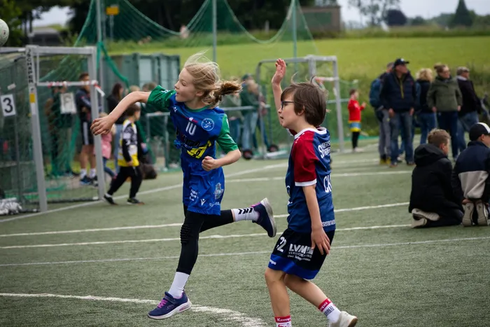 Zwei Kinder in Fußballtrikots springen auf einem Kunstrasenplatz. Das Mädchen in Blau und Grün ist in der Luft, der Junge in Blau und Rot versucht, den Ball zu spielen. Im Hintergrund stehen weitere Spieler und Zuschauer.