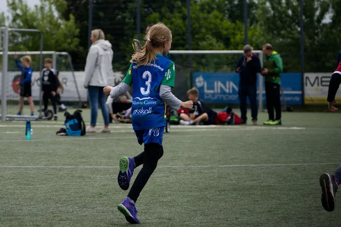 Ein junges Mädchen mit langen, blonden Haaren rennt auf einem Fußballfeld, während andere Spieler und Zuschauer im Hintergrund zu sehen sind. Sie trägt ein blaues Trikot mit der Nummer 3 und grüne Shorts.