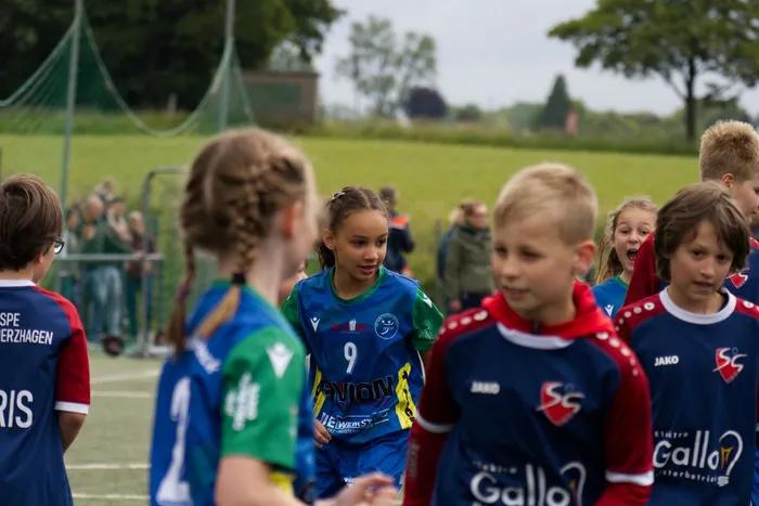 Eine junge Fußballspielerin mit der Nummer 9 auf dem Trikot geht auf dem Rasen zwischen anderen Kindern im Vordergrund vorbei. Im Hintergrund sind weitere Spieler und Zuschauer zu sehen.