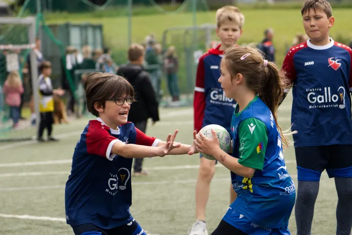 Zwei Kinder spielen auf einem Sportplatz Handball. Ein Mädchen in blauem Trikot wirft den Ball zu einem Jungen in rotem Trikot, während andere Kinder im Hintergrund zusehen.