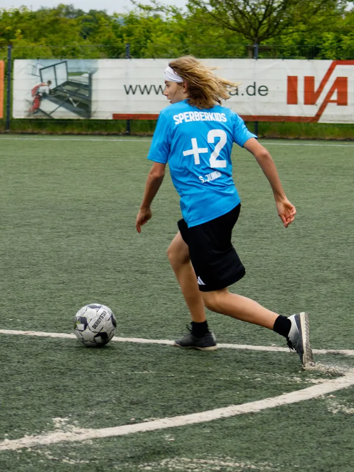 Ein Jugendlicher mit langen, blonden Haaren dribbelt einen Fußball auf einem grünen Kunstrasenplatz. Er trägt ein blaues Trikot mit der Nummer 2 und schwarze Shorts. Im Hintergrund ist eine Werbetafel zu sehen.