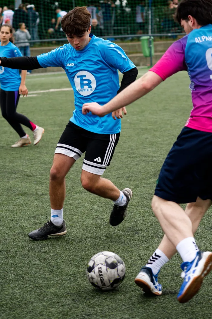 Ein junger Fußballspieler in blauem Trikot und schwarzen Shorts dribbelt den Ball auf einem grünen Kunstrasenplatz. Im Hintergrund sind weitere Spieler in bunten Trikots zu sehen.