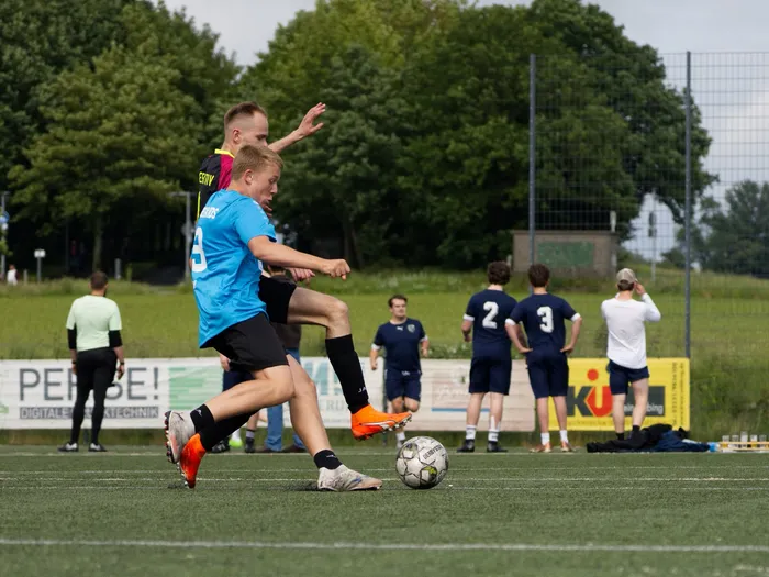 Zwei Fußballspieler kämpfen um den Ball auf einem grünen Rasenfeld. Im Hintergrund stehen weitere Spieler und Zuschauer an einem Zaun.