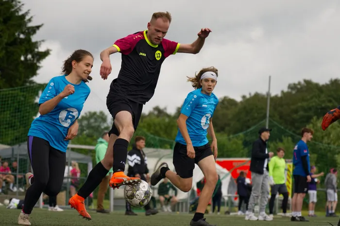 Ein Fußballspieler in rotem Trikot springt in der Luft, um den Ball zu treten, während zwei junge Spielerinnen in blauen Trikots versuchen, den Ball zu erobern. Im Hintergrund sind weitere Spieler und Zuschauer auf einem Fußballfeld zu sehen.