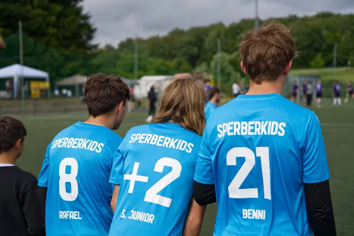 Drei junge Fußballspieler mit den Nummern 8, +2 und 21 stehen mit dem Rücken zur Kamera auf einem Fußballfeld. Auf ihren hellblauen Trikots steht 