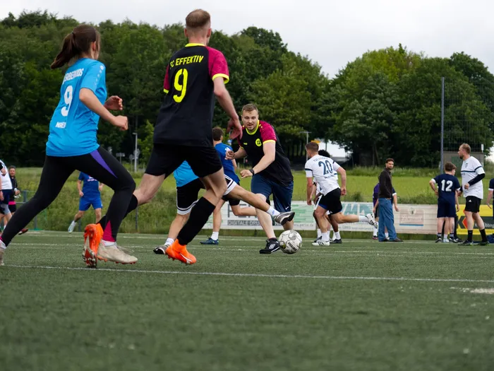 Fußballspieler in Aktion auf einem grünen Rasenfeld. Ein Spieler in schwarzem Trikot mit der Nummer 9 schießt auf das Tor, während andere Spieler um ihn herum stehen. Im Hintergrund sind weitere Spieler und Zuschauer zu sehen.
