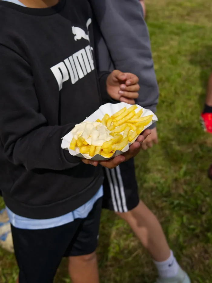 Eine Person hält eine Schale mit Pommes Frites in der Hand. Im Hintergrund sind weitere Personen und ein grüner Rasen zu sehen.
