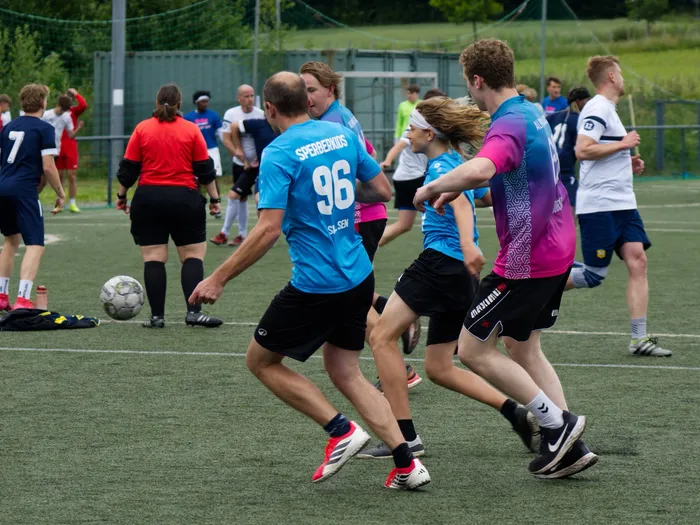 Fußballspieler in farbigen Trikots kämpfen um den Ball auf einem Kunstrasenplatz. Im Hintergrund sind weitere Spieler und Zuschauer zu sehen.