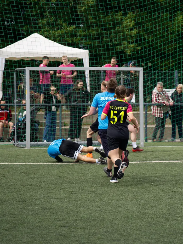 Ein Fußballspieler in schwarzer Hose und blauem Trikot rutscht auf dem grünen Kunstrasen, während ein Gegenspieler im gelben Trikot mit der Nummer 51 in der Nähe steht. Im Hintergrund stehen Zuschauer an einem Fußballfeld mit einem weißen Zelt.