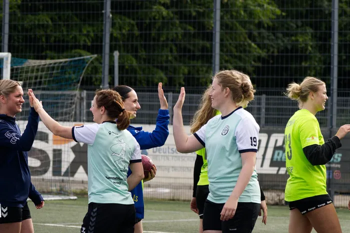 Vier junge Frauen in Sportkleidung stehen auf einem Rasenplatz und geben sich High-Fives. Im Hintergrund ist ein Zaun und Werbebanner zu sehen.