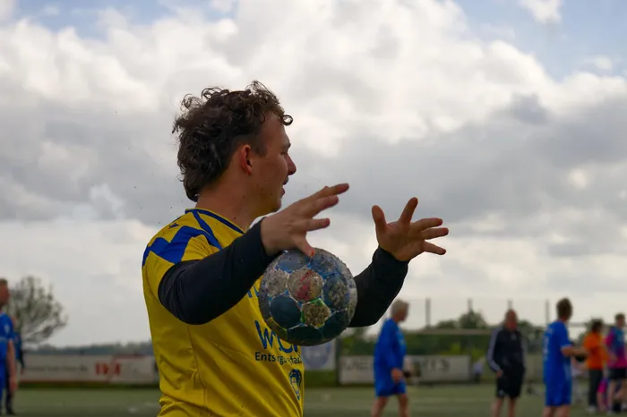 Ein Mann wirft einen Handball. Er trägt ein gelbes Trikot mit blauen Ärmeln und befindet sich auf einem Sportplatz vor Zuschauern.