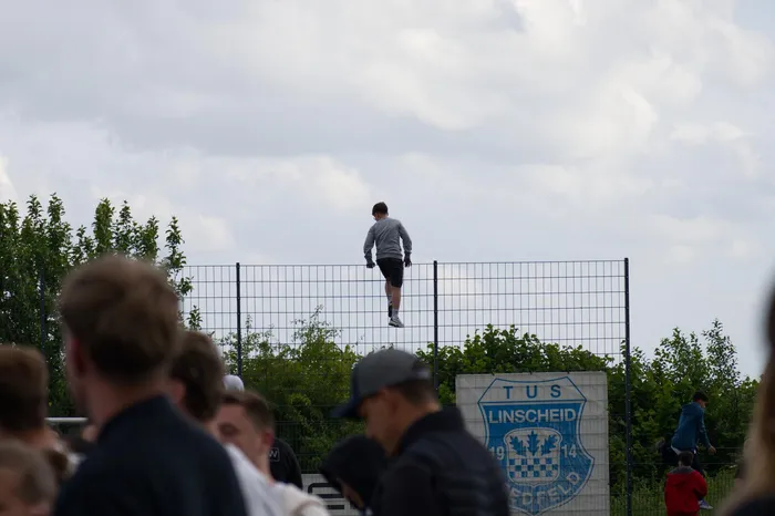 Ein Mensch steht auf einem Maschendrahtzaun vor Zuschauern und Bäumen. Im Hintergrund ist ein Schild mit der Aufschrift 