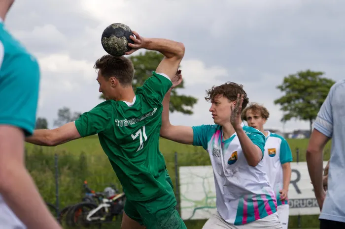 Ein Spieler in grünem Trikot wirft einen Handball, während ein Gegenspieler in blauem Trikot versucht, den Wurf zu blocken. Im Hintergrund sind weitere Spieler und Zuschauer zu sehen.