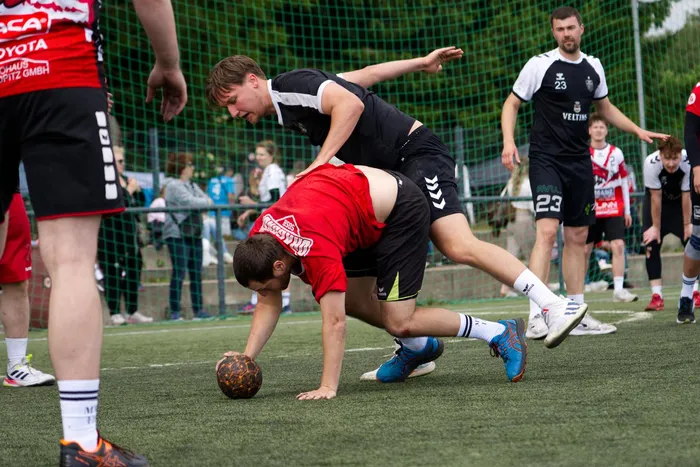 Ein Handballspieler in schwarzer und weißen Kleidung rutscht beim Versuch, den Ball zu erobern, an einem Spieler in rotem Trikot vorbei. Im Hintergrund sind weitere Spieler und Zuschauer auf dem Spielfeld zu sehen.