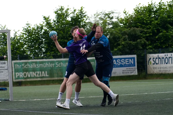 Ein Handballspieler in violettem Trikot wirft den Ball, während ein Gegenspieler in blauem Trikot ihn blockt. Im Hintergrund ist ein Handballfeld und Werbebanner zu sehen.