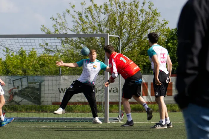 Ein Handballtorwart in weiß-blauem Trikot springt zur Abwehr, während ein Angreifer im roten Trikot zum Wurf ausholt. Im Hintergrund stehen weitere Spieler auf dem Spielfeld.