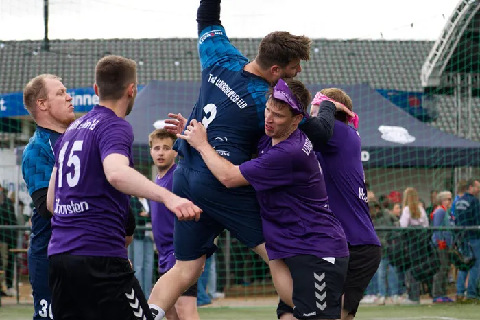 Spieler des Handballteams feiern auf dem Spielfeld, indem sie ihren Torwart in die Höhe werfen. Die Spieler tragen violette Trikots mit der Aufschrift 'Hosten' und 'Hummel'. Im Hintergrund sind weitere Zuschauer und ein Stadion zu sehen.