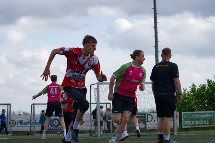 Ein Fußballspieler in rotem und weißem Trikot rennt auf das Tor zu, während ein anderer Spieler in pinkem Trikot ihm folgt. Im Hintergrund steht ein Schiedsrichter.