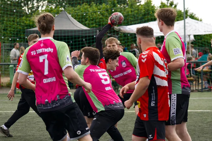 Handballspieler in farbigen Trikots kämpfen um den Ball auf einem Outdoor-Spielfeld. Im Hintergrund ein graues Zeltdach und Zuschauer.