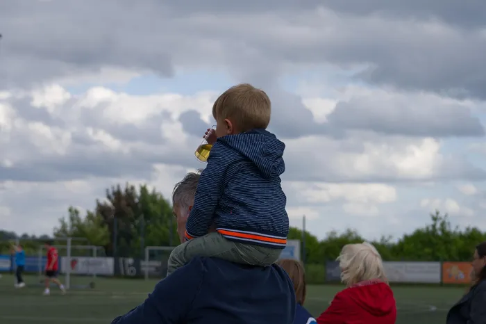 Ein kleines Kind sitzt auf den Schultern einer Person und hält ein Getränk in der Hand. Im Hintergrund ist ein Handballfeld und Zuschauer zu sehen.