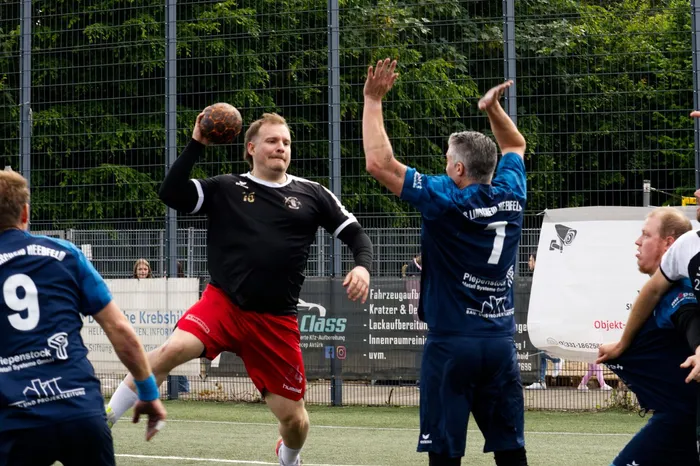 Ein Handballspieler in schwarzem Trikot und roten Shorts wirft den Ball während eines Spiels auf Kuhlenhagen. Ein Gegenspieler in blauem Trikot versucht, den Wurf zu blocken.