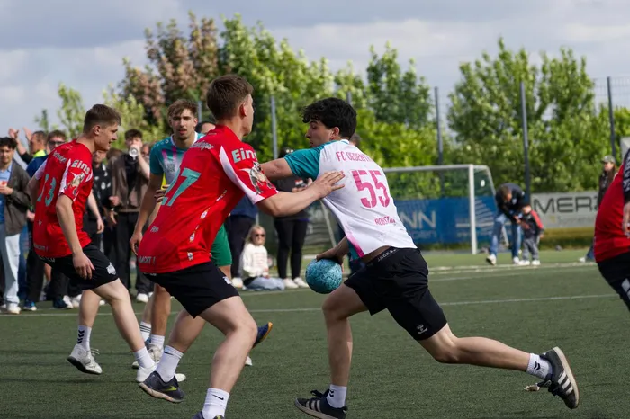 Handballspieler kämpfen um den Ball auf dem Spielfeld, während Zuschauer im Hintergrund zusehen. Ein Spieler in schwarzer Hose und weißem Trikot mit der Nummer 55 versucht, den Ball zu erobern.