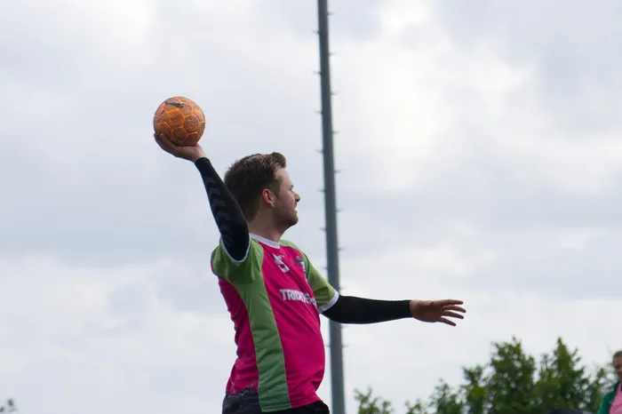 Ein Mann wirft einen Handball hoch in die Luft. Er trägt ein pink-grünes Trikot und Armstulpen.