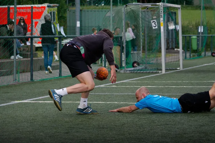 Ein Spieler in schwarzer Sportkleidung und ein Spieler in blauem Trikot kämpfen um einen Ball auf einem grünen Handballfeld. Im Hintergrund steht ein rotes Werbebanner und ein Tor.