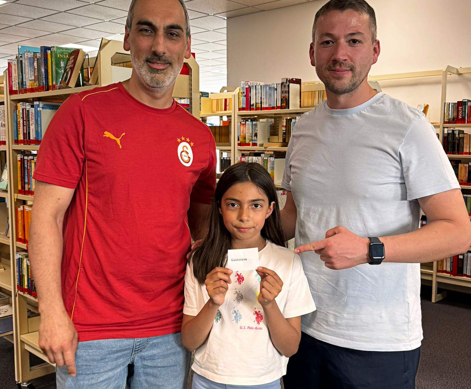 Ein Mann links im Galatasaray-Trikot, ein Mann rechts in einem weißen Hemd und ein Mädchen in der Mitte halten ein Los mit der Aufschrift „Galatasaray“ vor sich in einer Bibliothek.