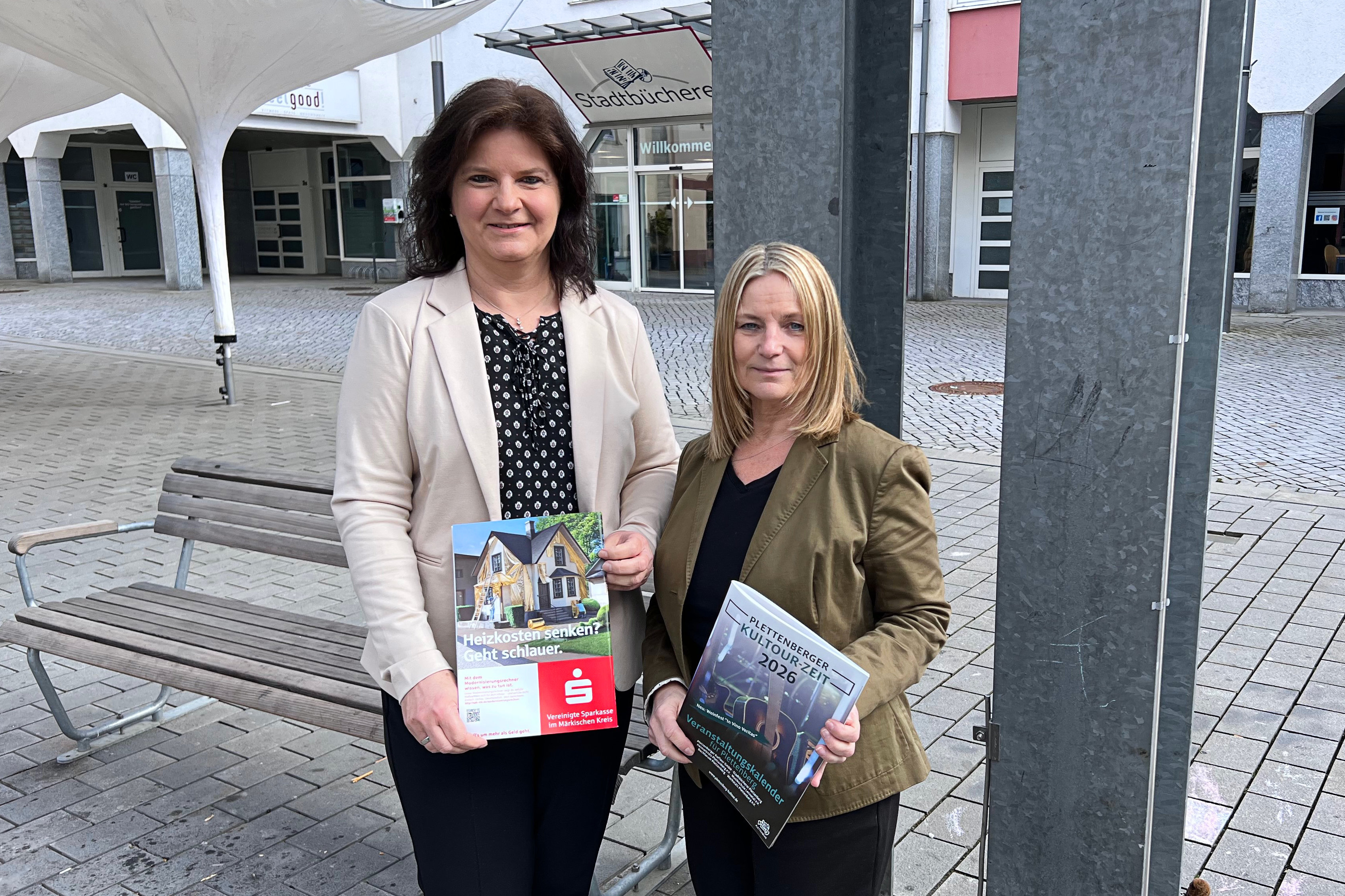 Zwei Frauen stehen lächelnd vor einer Stadtbücherei und halten Informationsmaterialien in den Händen: Sylvia Eick (rechts) präsentiert die Broschüre KulTourZeit 2026, Manuela Keiderling hält eine Broschüre zum Thema Heizkostensenkung.