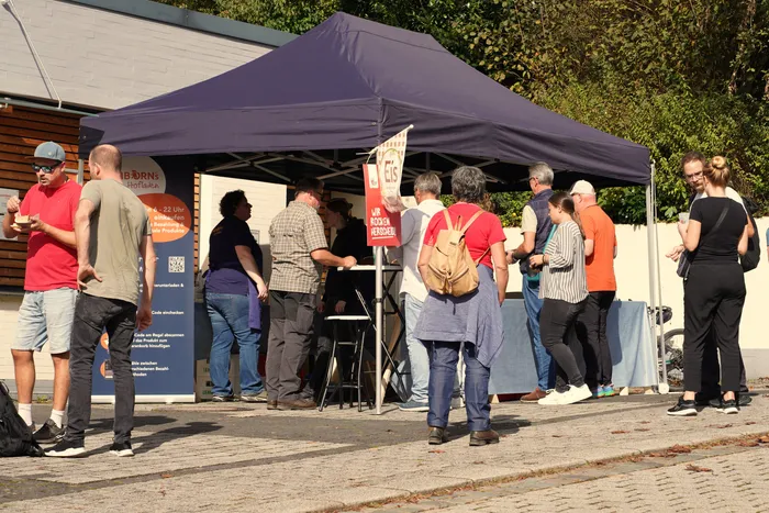 Menschen an einem blauen Stand unter einem Zelt bei der kulinarischen Wanderung in Herscheid.