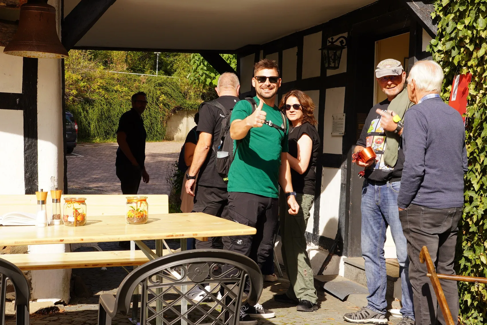 Ein Mann im grünen T-Shirt gibt der Kamera einen Daumen nach oben, während er mit einer Gruppe von Menschen vor einem Fachwerkhaus steht.