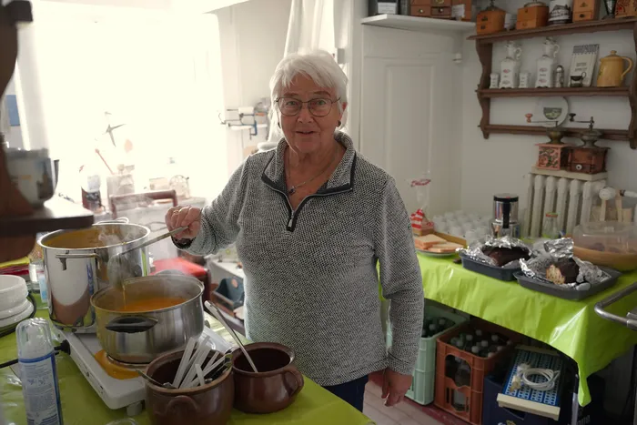 Eine ältere Frau mit Brille rührt mit einer Kelle in einem großen Topf mit gelber Suppe.