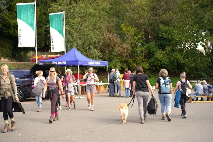 Wandernde Personen gehen an einem blauen Zelt und grünen Fahnen vorbei, ein Hund läuft auf dem Weg.