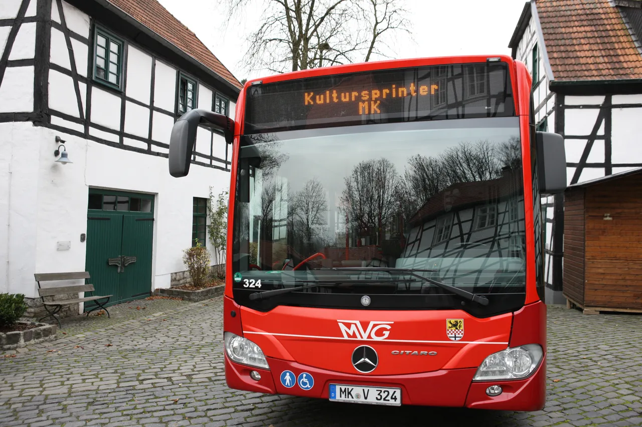Roter Bus mit der Aufschrift „Kultursprinter MK“ steht vor einem Fachwerkhause in einem Pflasterhof.