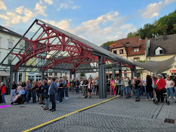 Menschenmenge unter einer markanten roten Stahlträger-Konstruktion auf einem gepflasterten Platz.