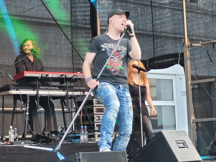 Ein Sänger der Coverband Rockwood singt auf einer Bühne in ein Mikrofon, im Hintergrund sind ein Keyboarder und eine weitere Bandmitglied zu sehen.