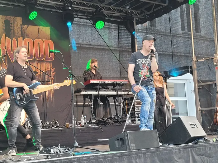 Die Coverband Rockwood tritt auf einer Bühne auf, mit einem Bassisten, einem Keyboarder, einem Sänger am Mikrofon und einer Sängerin.