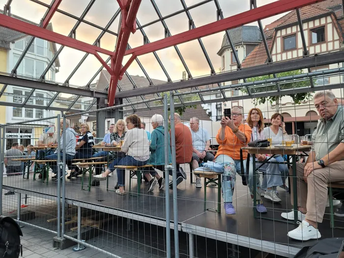 Menschen sitzen an Tischen auf einer Bühne unter einer roten Metallkonstruktion mit Glasdach.