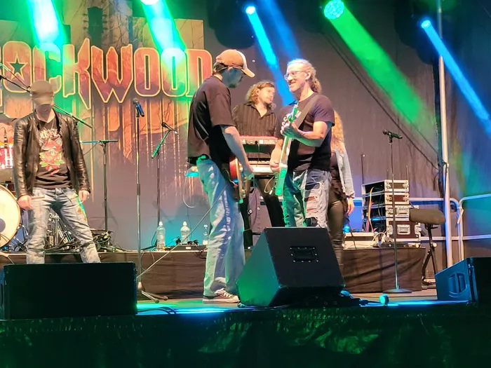 Mitglieder der Coverband Rockwood spielen auf einer Bühne vor einem großen Schriftzug unter grünem und blauem Scheinwerferlicht.