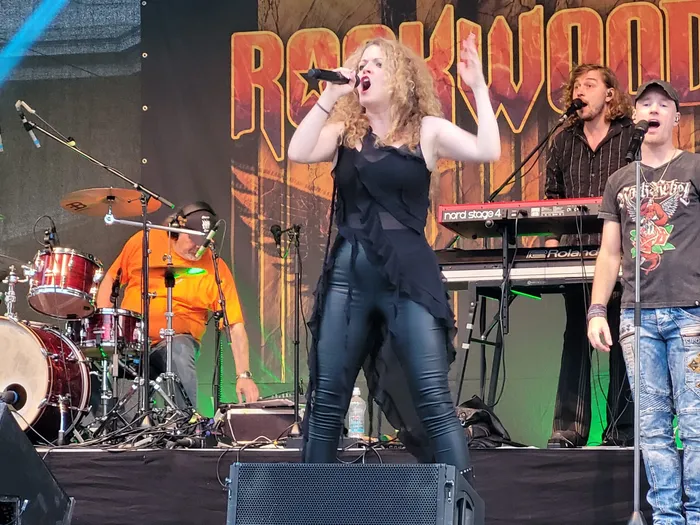 Eine Sängerin singt auf einer Bühne vor einem Rockwood-Banner, begleitet von einem Schlagzeuger und weiteren Bandmitgliedern.