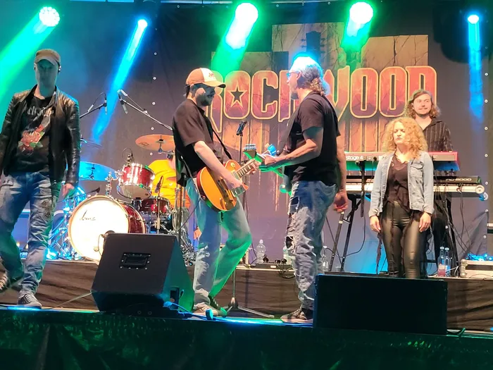 Die Coverband Rockwood spielt live auf einer Bühne mit farbiger Beleuchtung vor einem großen „Rockwood“-Schriftzug.