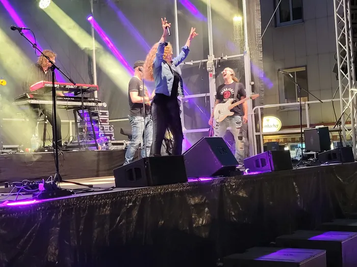 Die Band Rockwood tritt auf einer Bühne unter violetten Scheinwerfern auf, wobei die Sängerin im Zentrum die Arme hebt.