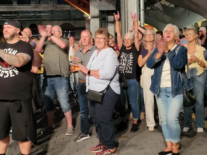 Ein Publikum klatscht, hält Getränke und zeigt Rock-Handzeichen bei einem Konzert.