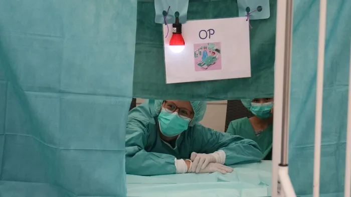 Eine Person in medizinischer Schutzkleidung und Maske blickt durch eine Öffnung in einem türkisfarbenen Vorhang, über der ein Schild mit der Aufschrift „OP“ hängt.