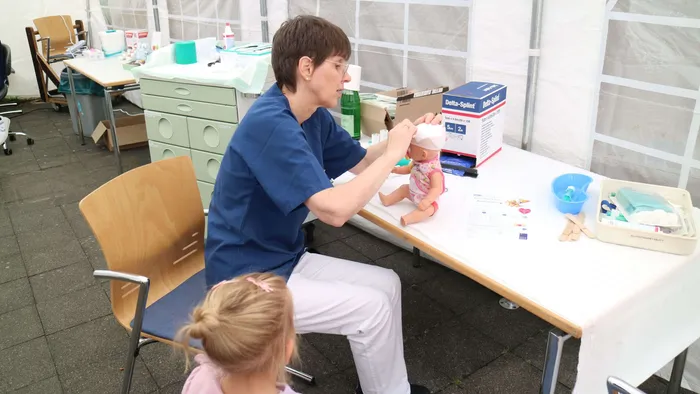 Eine Frau in medizinischer Kleidung legt einer Puppe an einem Tisch mit medizinischem Zubehör einen Verband an.