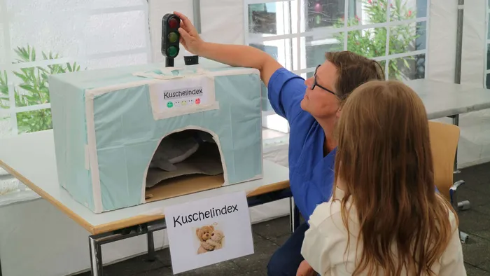 Eine Frau in blauer Kleidung bedient eine Ampel neben einer hellblauen Box mit der Aufschrift „Kuschelindex“, in der ein weißes Kuscheltier liegt.