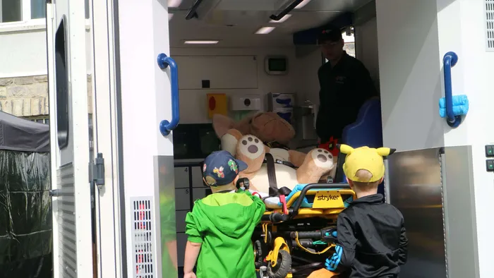 Zwei Kinder blicken in einen Rettungswagen, in dem ein großes Kuscheltier auf einer Trage liegt.