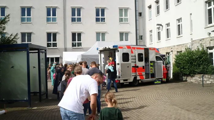 Menschenmenge vor einem weißen Zelt und einem Rettungswagen in einem gepflasterten Innenhof vor einem weißen Gebäude.